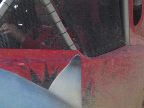 1990 Ford Bronco - Tight detail of scuffed red and silver bodywork on an off-road race buggy, showing the roll cage tubing and mesh window panels.