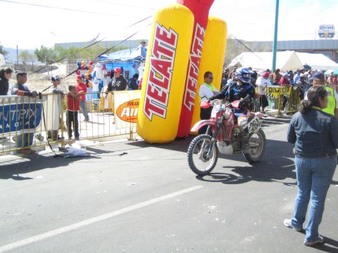 1990 Ford Bronco - rider on a red off-road motorcycle waits at the start line next to a yellow inflatable Tecate banner with spectators behind barriers.