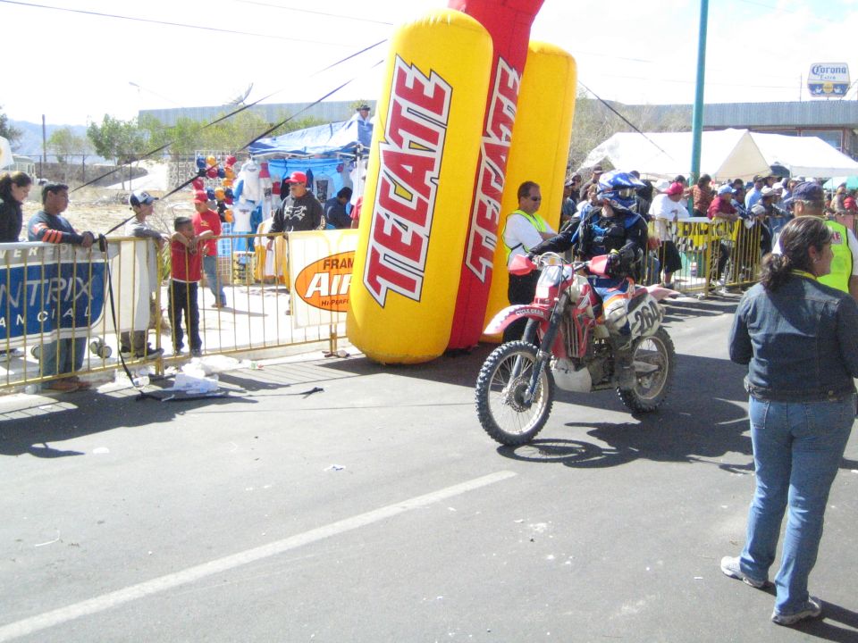 Off-road motorcycle rider at a race start line beside a yellow inflatable Tecate arch and crowd