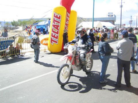 1990 Ford Bronco - helmeted dirt-bike rider in racing gear pauses on the road beside a yellow Tecate inflatable banner surrounded by spectators.