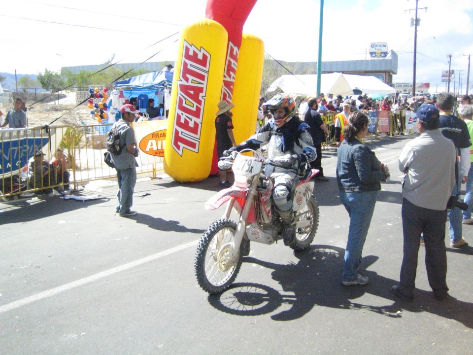 Motorcyclist in racing gear on a dirt bike beside a Tecate inflatable banner with crowds