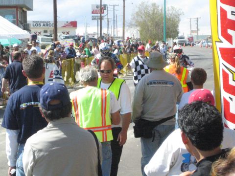 1990 Ford Bronco - crowd of spectators and officials lines a street race course, with a man in a yellow safety vest and a checkered flag visible among them.