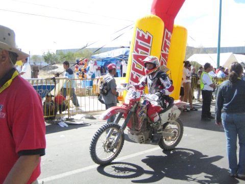 1990 Ford Bronco - rider on a red number 504 off-road motorcycle pauses beside a yellow Tecate inflatable banner with onlookers around the course.