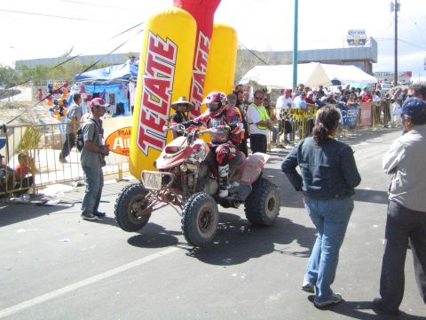 1990 Ford Bronco - Two riders on a red racing ATV quad pause beside a yellow Tecate inflatable banner as spectators watch from the roadside.