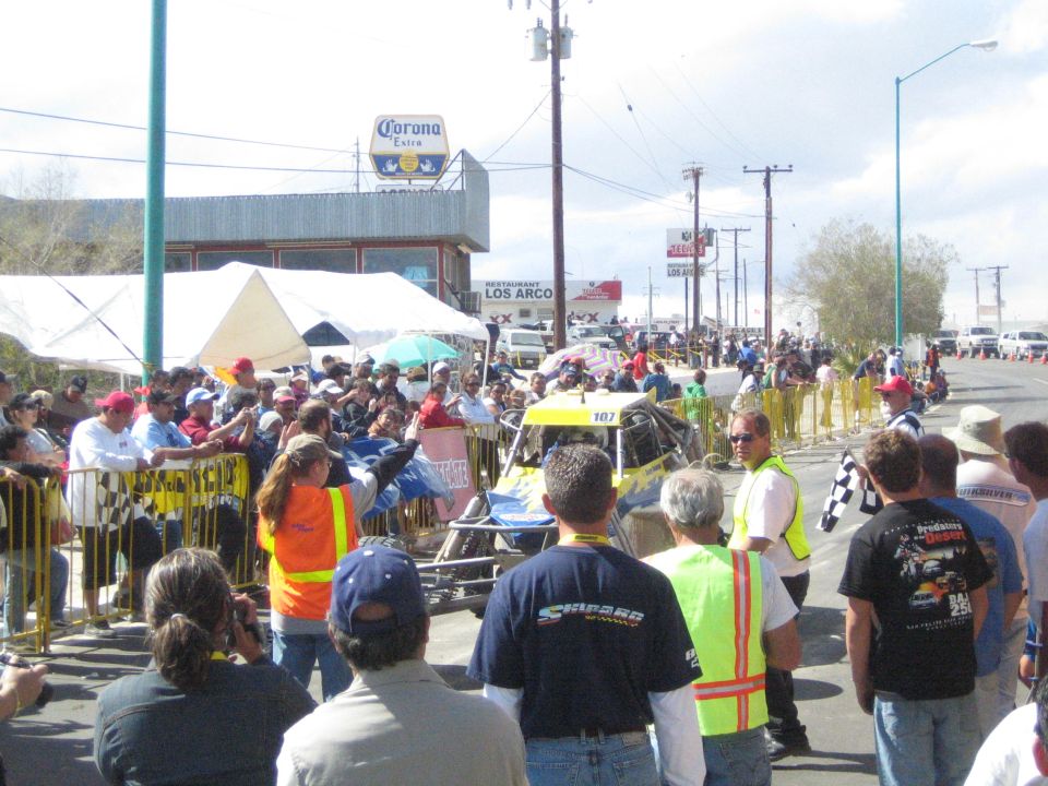 Crowd watching an off-road buggy on a street under a Corona sign and Los Arcos restaurant