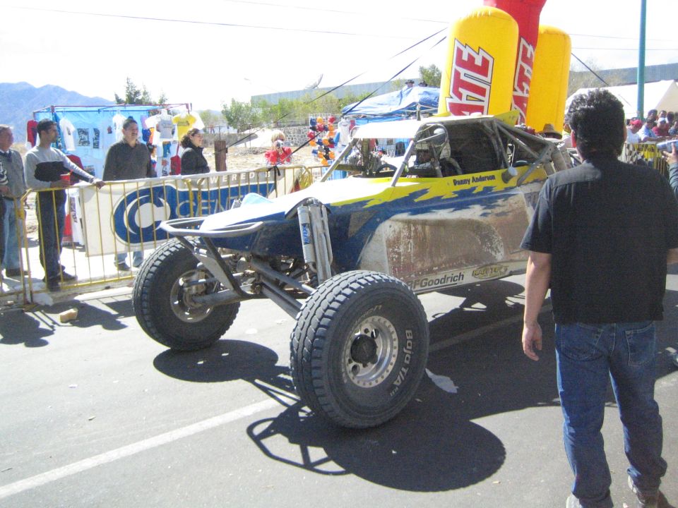 Blue and white off-road race buggy parked on a street with spectators and Tecate inflatable