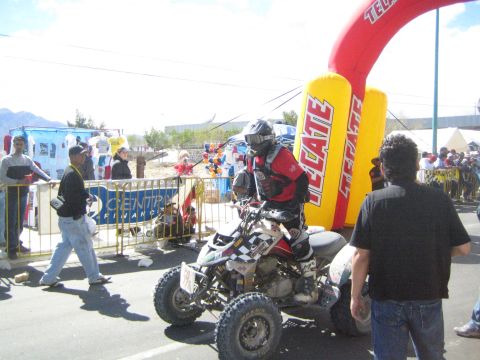1990 Ford Bronco - helmeted rider sits on a racing ATV quad next to a yellow Tecate inflatable arch with spectators behind the barriers.
