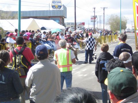 1990 Ford Bronco - Spectators and a man in a yellow vest line a street race course where a rider passes, with a checkered flag and Corona sign in view.
