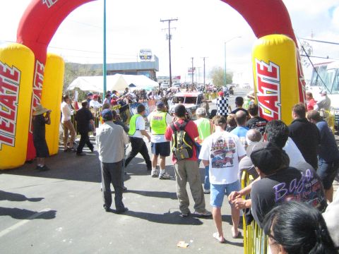 1990 Ford Bronco - street race start line framed by twin yellow Tecate inflatable arches, crowded with spectators and a checkered flag.