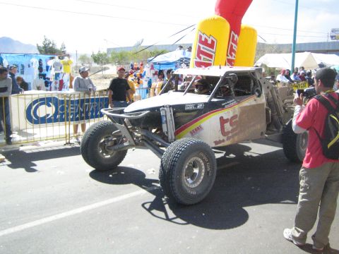 1990 Ford Bronco - white and silver tube-frame off-road race buggy moves along a street as a photographer and crowd watch near a Tecate inflatable.