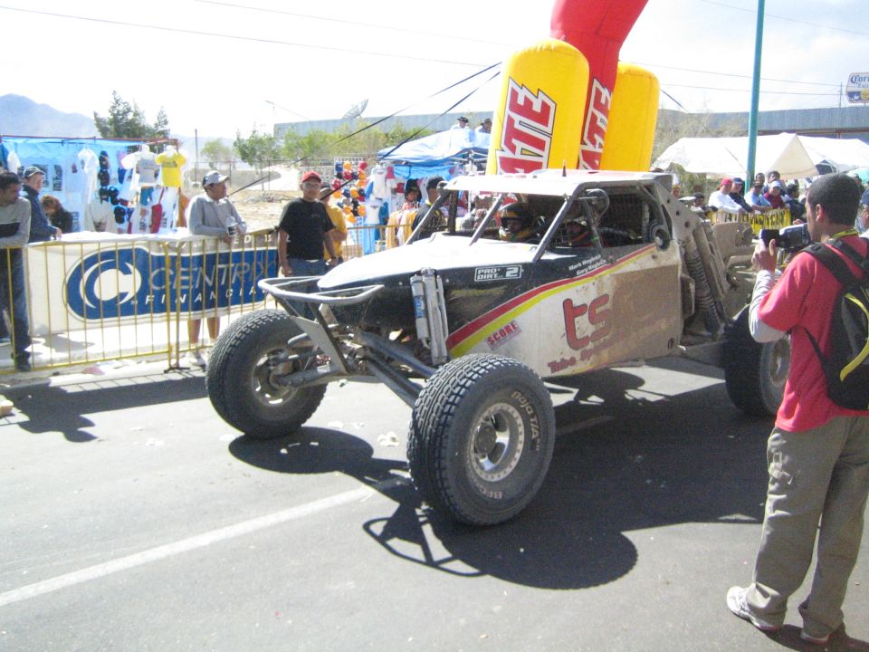 White and silver off-road race buggy on a street with photographer and crowd, Tecate inflatable