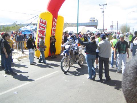 1990 Ford Bronco - helmeted dirt-bike rider raises a hand to greet someone beside a yellow Tecate inflatable banner amid a crowd of spectators.