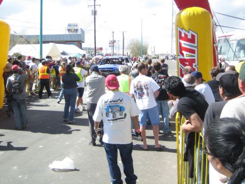 1990 Ford Bronco - Spectators behind yellow barriers watch a dark pickup truck on a street course, with a Corona sign and Tecate banner in the background.