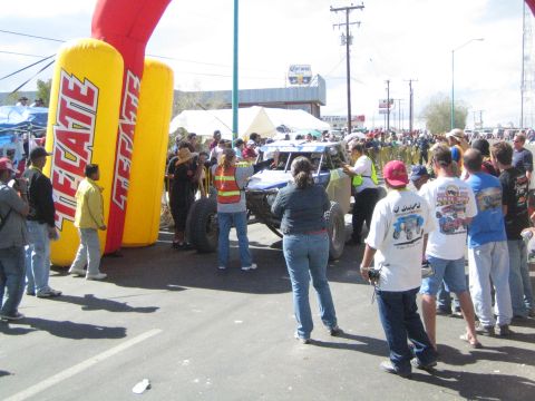 1990 Ford Bronco - blue and white off-road race buggy is surrounded by spectators and officials at the start line beside a yellow Tecate inflatable.