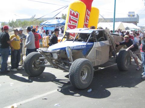 1990 Ford Bronco - blue and silver off-road race buggy with BFGoodrich markings is pushed by a man along a street as spectators look on.
