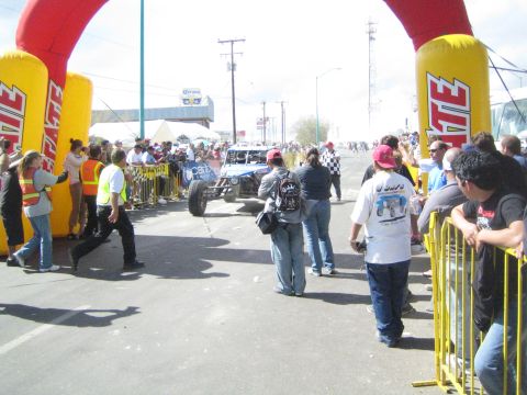 1990 Ford Bronco - blue off-road race buggy travels down a street between twin yellow Tecate inflatable arches lined with a large crowd.