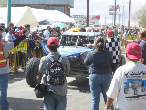 1990 Ford Bronco - blue off-road race buggy is surrounded by a crowd at the start area, with one man carrying a spare tire and a Los Arcos sign behind.
