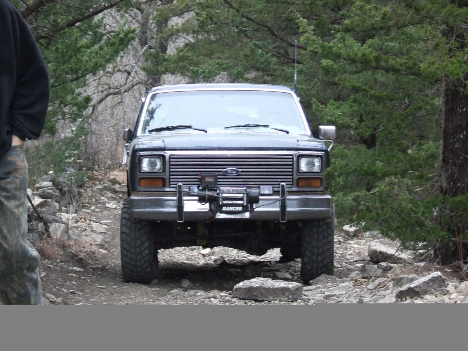 Front view of a dark-colored Ford Bronco on a rocky, wooded trail. It has a winch mounted on the front bumper