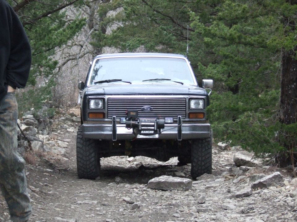 Front view of a 1986 Ford Bronco with a winch on the bumper, driving on a rocky trail