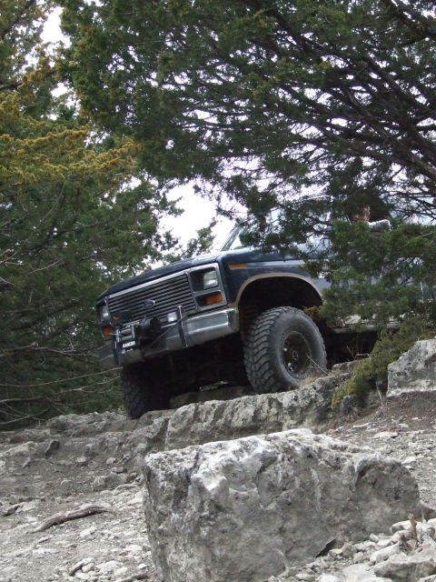 1986 Ford Bronco - This Ford Bronco is navigating a rocky, off-road path with dense trees providing shade.