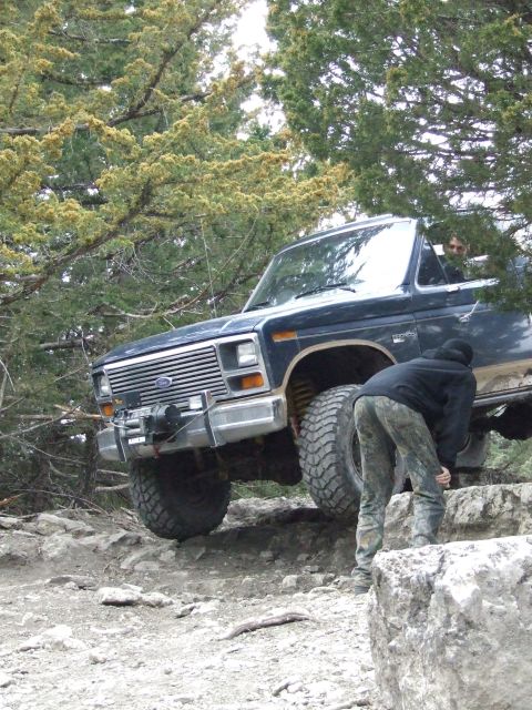 1986 Ford Bronco - A dark blue 1986 Ford Bronco navigates a rocky trail, with a person attending to the front tire.