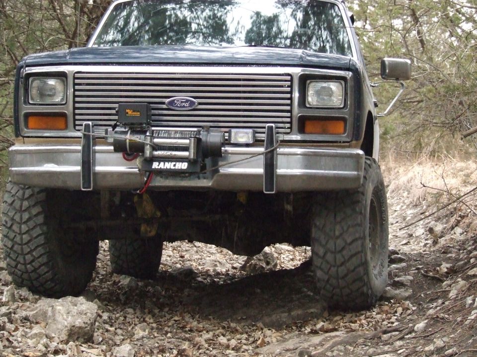 Front view of a dark blue Ford Bronco with a winch mounted on the bumper, driving on a rocky trail
