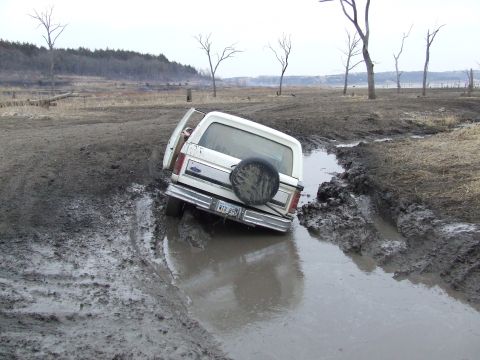 1986 Ford Bronco - A white Ford Bronco is stuck in a muddy rut with its driver's door open.