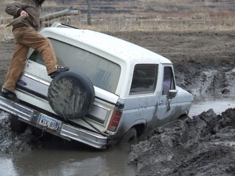 1986 Ford Bronco - A 1986 Ford Bronco is stuck deep in mud, with a person standing on its rear bumper.