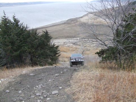 1986 Ford Bronco - A Ford Bronco navigates a rough, unpaved road with a large body of water in the background.