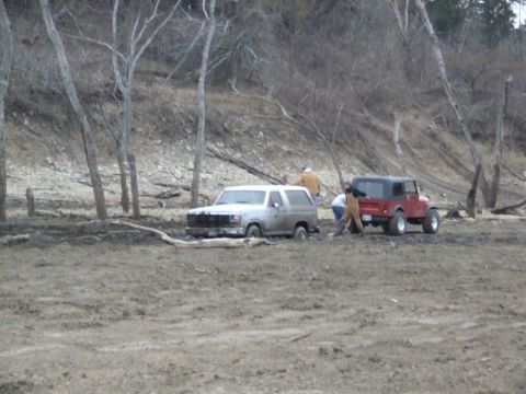 1986 Ford Bronco - Two off-road vehicles are stuck in thick mud near a wooded area, with people attempting to assist.