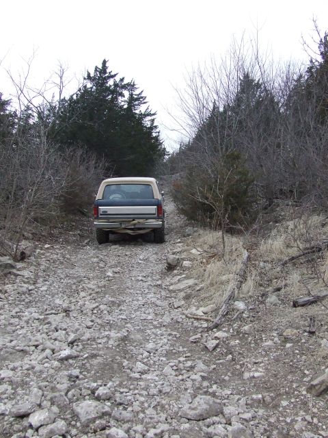 1986 Ford Bronco - A tan and blue Ford Bronco drives up a rocky, unpaved road surrounded by trees.
