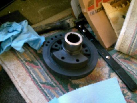 1986 Ford Bronco - A crankshaft pulley sits on a patterned rug next to blue shop towels and a cardboard box.