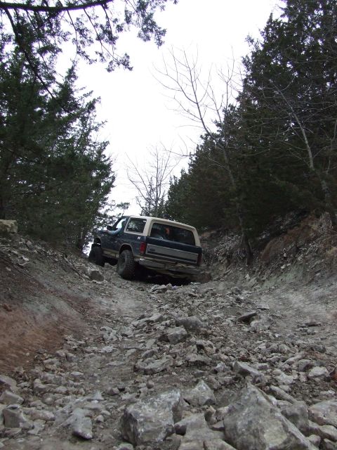 1986 Ford Bronco - A 1986 Ford Bronco navigates a challenging rocky incline surrounded by trees.