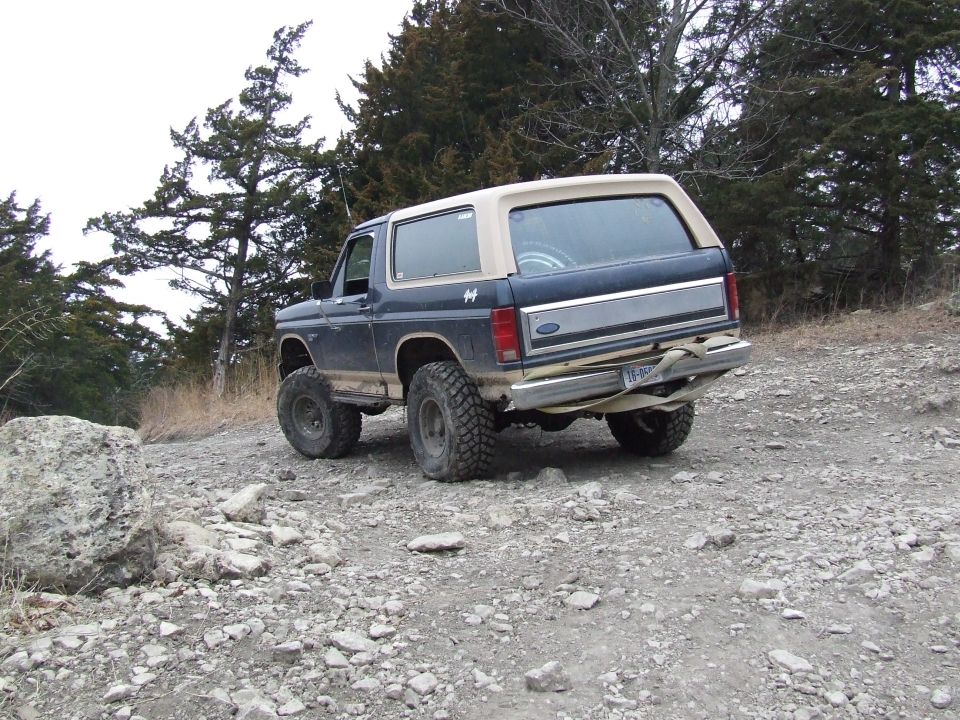 Ford Bronco on a rocky trail, rear view. It has a tan camper shell and big off-road tires