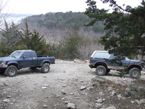 1986 Ford Bronco - A 1986 Ford Bronco and a pickup truck are parked on a rocky trail with trees and hills in the background.