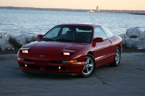 1993 Ford Probe - A red 1993 Ford Probe is parked near the water with a city skyline in the distance.