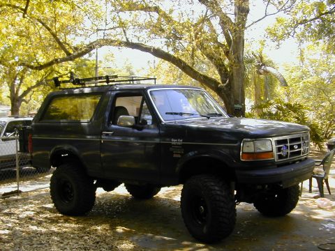 1993 Ford Bronco - A dark green two-door Ford Bronco full-size SUV with a lift kit, black steel wheels, and large mud-terrain tires, viewed from the front-rig…