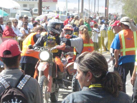 1990 Ford Bronco - helmeted off-road motorcycle racer waits at a packed start line as spectators and orange-vested officials crowd around.