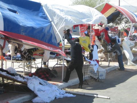 1990 Ford Bronco - vendor arranges canopy tents and racks of clothing at an outdoor market under bright sun.