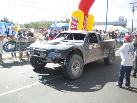 1990 Ford Bronco - dirt-covered off-road race pickup truck rolls under a yellow Tecate inflatable arch as a crowd watches behind barriers.