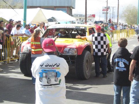 1990 Ford Bronco - red and yellow off-road race truck passes officials waving a checkered flag in front of a roadside crowd.