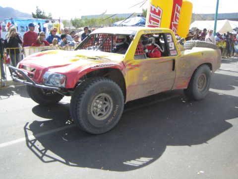 1990 Ford Bronco - weathered red and yellow trophy-style off-road race truck, number 39, sits on the paved course before a crowd.