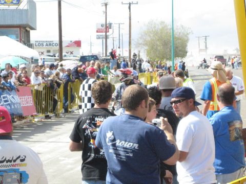 1990 Ford Bronco - dense crowd in graphic T-shirts and caps lines a sunny roadside, watching an off-road race near a Los Arcos restaurant.