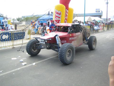 Red open tube-frame off-road race buggy on pavement under Tecate arch