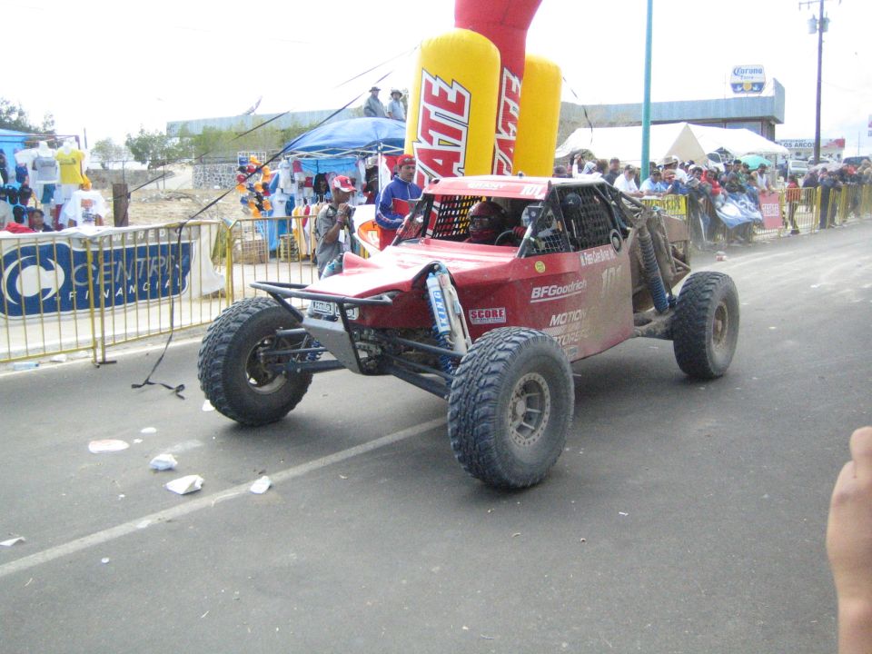 Red open tube-frame off-road race buggy on pavement under Tecate arch