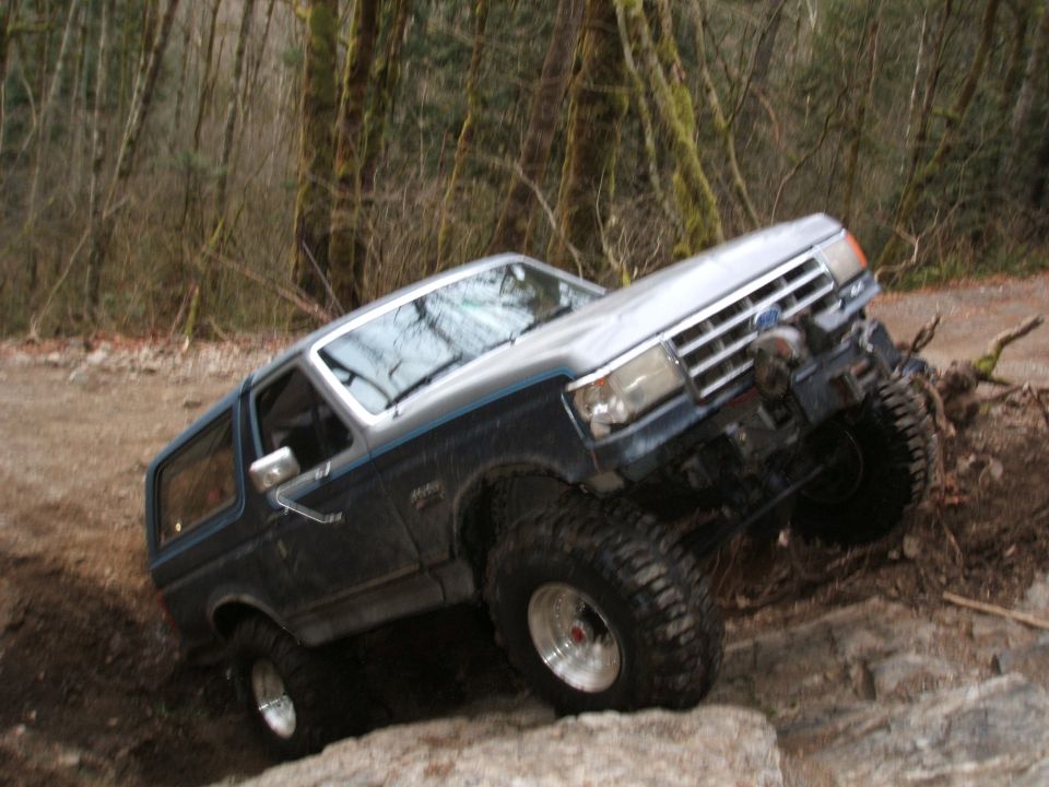 Blue and silver lifted Ford Bronco crawling over rocks on a wooded off-road trail
