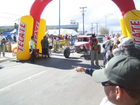 Spectator pointing toward a red race buggy under a Tecate arch on a street