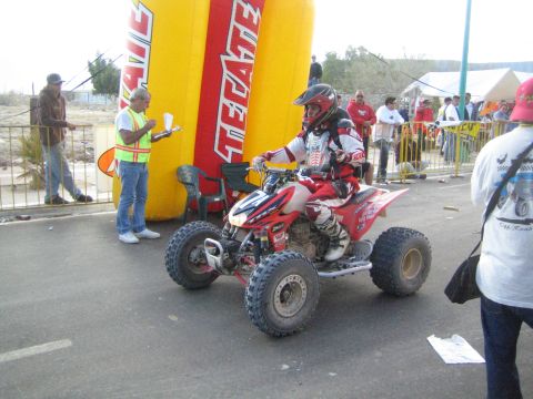Rider on a red and white racing ATV beside a Tecate arch on a street