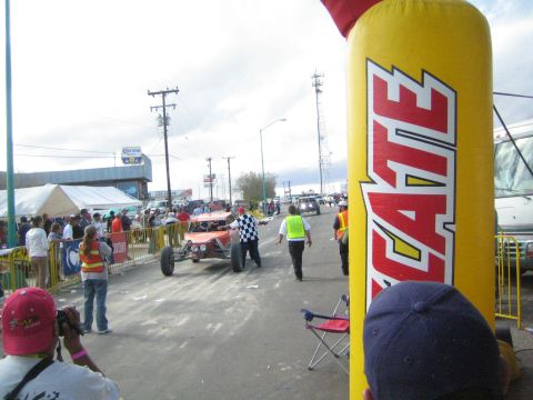 Orange race buggy on a street beside a large yellow Tecate arch