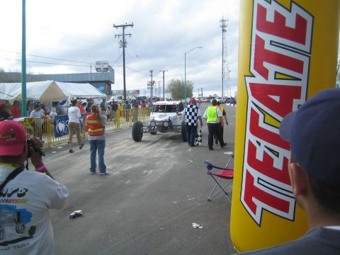 Off-road race buggy at staging area with spectators and tall yellow Tecate inflatable column